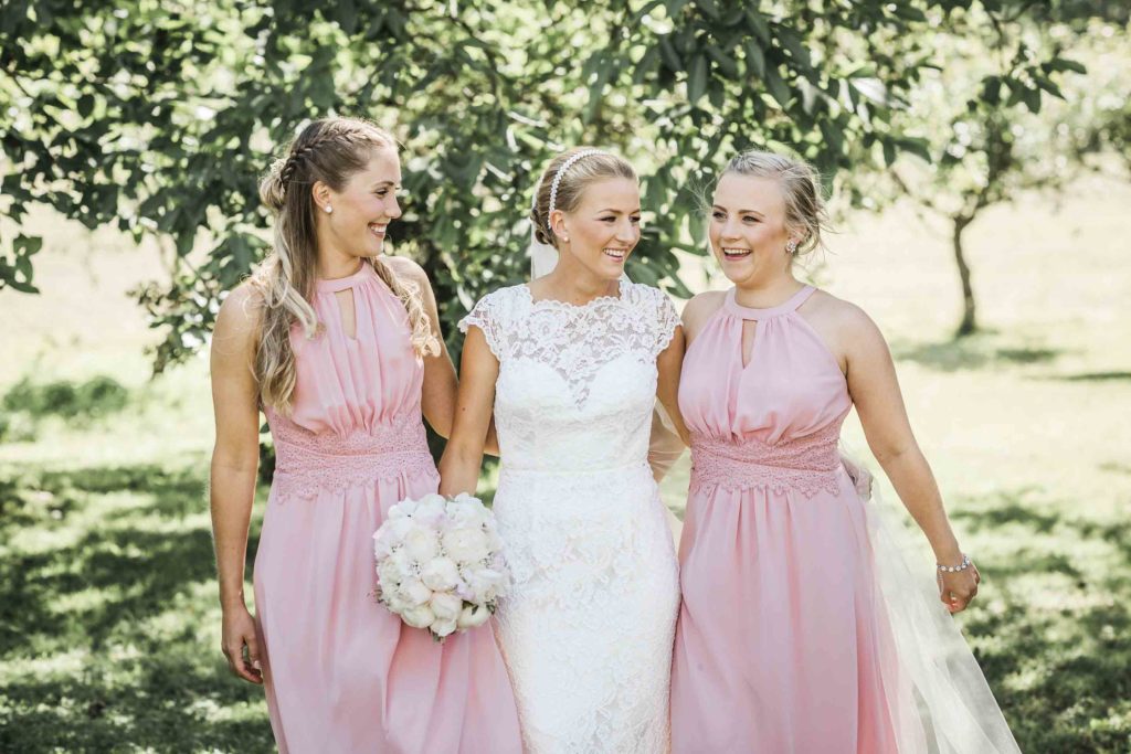 Brud i hvit blondekjole med brudebukett i peoner og oppsatt hår. Forlovere med rosa kjoler.
